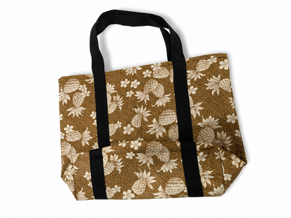 A brown canvas tote bag with a cream pineapple and floral print, featuring two black webbed handles, shown on a plain white background.